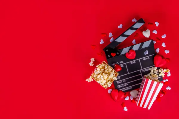 Romantic date on Valentine\'s Day February 14. Love movie marathon, date at cinema theater. Clapperboard, popcorn buckets, heart decor and chocolates on red background flat lay copy space