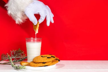 Noel Baba 'nın eli, geleneksel Noel kurabiyesi atıştırmalığıyla süt bardağıyla. Santa Dunking Cookie, parlak kırmızı ve beyaz ahşap arka planda. Mutlu Noeller ve mutlu yıllar tebrik kartı.
