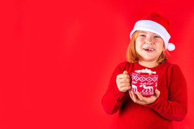 Funny caucasian girl with cup of Christmas cocoa. Happy cute european blonde girl in red sweater and Santa hat hold and drink cup of hot chocolate with marshmallow, on red background