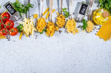 Beyaz masa arkasında çeşitli kurutulmuş spagetti, makarna ve kaşıklarla hazırlanmış makarna. Farklı şekillerde çiğ İtalyan makarnası, otlar, zeytinyağı, domatesler, baharatlar.