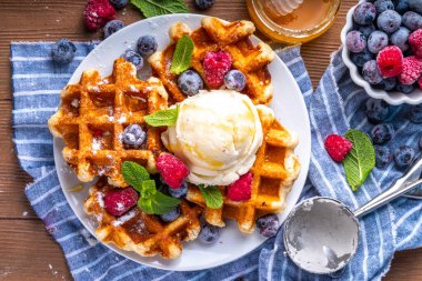 Tasty Homemade Belgian waffles with raspberries, blueberries, vanilla ice cream and syrup or honey, sweet summer healthy breakfast, light wooden background copy space 