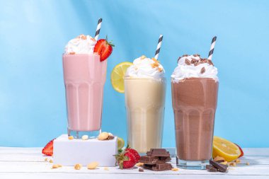Yaz tazeleyici milkshake seti ya da klasik lezzetli tadı olan meyve suları vanilya limonu, çilek, çikolata, krem şanti ve malzemelerle beyaz, açık mavi, güneşli bir arka planda. 