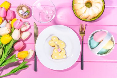 Şirin Paskalya sofrası, bahar tatili tabağı, cam, çatal bıçak takımı, lale çiçeği buketi, Paskalya dekorasyon sembolleri ve ikramlar, pembe ahşap parlak masa, üst görünüm kopya alanı.