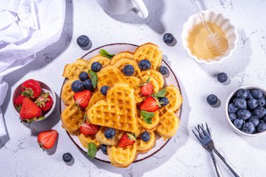 Çilekli, yaban mersinli ve ballı Belçika waffle 'ları, beyaz mutfak masalarında ev yapımı kalp şeklinde waffle' lar.