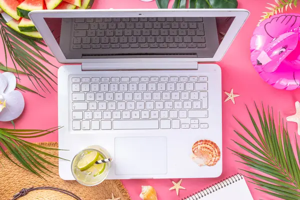 Summer office, working background, blogger workplace flat lay. White laptop keyboard with notebook, straw hat, tropical decor and leaves, cold cocktail glass on high-colored sunny pink background
