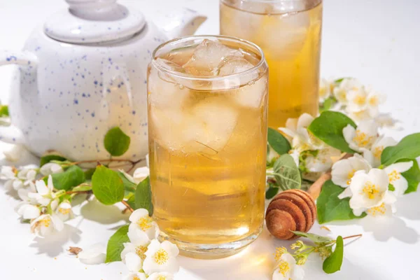 Organic herbal health summer cold drink. Jasmine flowers green tea, in glass, with ice cubes, teapot, on white table, with blooming jasmine flowers