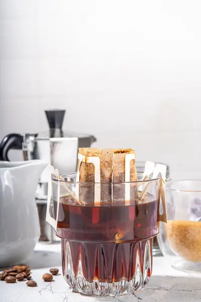 Trendy instant brewed drip coffee, cold brew coffee. Drip filter coffee bag in glass cup on white table, with brown sugar, coffee beans, pot with hot water