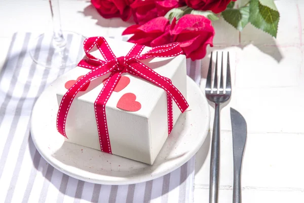 Valentine day table setting with plate, gift box with festive red ribbon, wine glass, fork and knife, red roses flowers bouquet white tiled table flatlay copy space