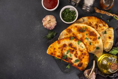 Cheburekler, börekler. Kızarmış turtalar, et ve baharatlı, otlar ve domates sosuyla birlikte siyah beton bir arka planda.
