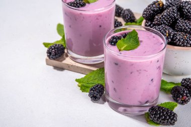 İki bardak böğürtlenli smoothie, yoğurt veya taze böğürtlenli milkshake, beyaz masa arkasında nane yapraklı fotokopi alanı.