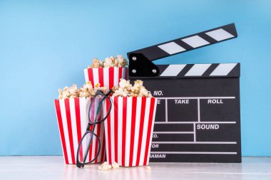 Sinema, film ve film izleme gecesi konsepti. Alkış tahtası, çizgili patlamış mısır kovası kutuları, açık mavi arka planda gözlük, kopyalama alanı