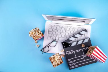 Sinema, film ve film izleme gecesi konsepti. Alkış tahtası, çizgili patlamış mısır kovası kutuları, açık mavi arka planda gözlüklü ve beyaz dizüstü bilgisayarlı, boşluğu kopyala