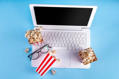 Sinema, film ve film izleme gecesi konsepti. Alkış tahtası, çizgili patlamış mısır kovası kutuları, açık mavi arka planda gözlüklü ve beyaz dizüstü bilgisayarlı, boşluğu kopyala