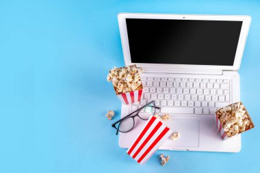 Sinema, film ve film izleme gecesi konsepti. Alkış tahtası, çizgili patlamış mısır kovası kutuları, açık mavi arka planda gözlüklü ve beyaz dizüstü bilgisayarlı, boşluğu kopyala