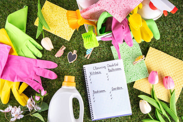 Весенняя уборка фона. Комплект чистящих средств, бутылок, аксессуаров и вещей с весенними цветами в корзине на белом деревянном зеленом саду