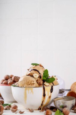 Fındıklı çikolatalı dondurma. Fındıklı dondurma kepçeli büyük kase porsiyonu, çikolata sosu ve mutfak masasının arkasında bir sürü fındık. 