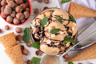 Fındıklı çikolatalı dondurma. Fındıklı dondurma kepçeli büyük kase porsiyonu, çikolata sosu ve mutfak masasının arkasında bir sürü fındık. 