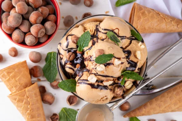 Fındıklı çikolatalı dondurma. Fındıklı dondurma kepçeli büyük kase porsiyonu, çikolata sosu ve mutfak masasının arkasında bir sürü fındık. 