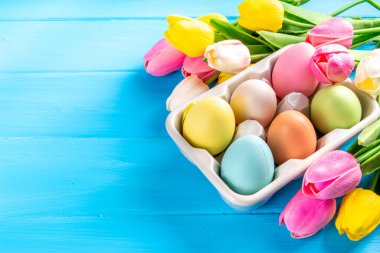 Simple minimal Easter greeting card background, colorful pastel Easter eggs with tulip flower bouquet, on light blue wooden plank background top view copy space