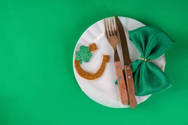 Saint Patrick masa takımı çatal bıçak, bıçak ve çatal, Patrick günü dekorasyonu, bozuk paralar, leprikon şapkası, gözlükler, şanslı at nalı, konfeti, yeşil arka planda boncuklar.