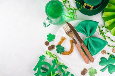 Saint Patrick masa takımı çatal bıçak, bıçak ve çatal, Patrick günü dekorasyonu, bozuk paralar, leprikon şapkası, gözlükler, şanslı at nalı, konfeti, yeşil arka planda boncuklar.
