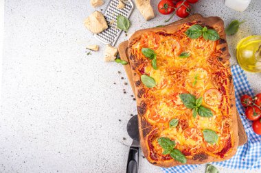 Domates ve fesleğenli ev yapımı margarita pizza, malzemeler ve arka planda beyaz mutfak masasında zeytinyağı.