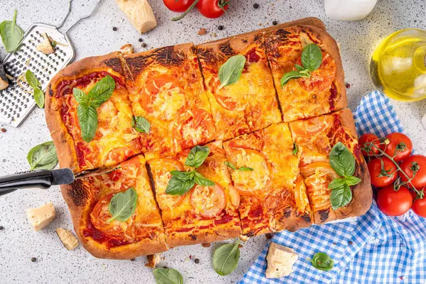 Rectangular homemade margarita pizza with tomatoes and basil, with ingredients and olive oil on white kitchen table background