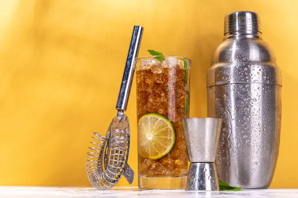 Cuba libre or long island summer cocktail, iced cola with lime and mint. Iced sweet long alcohol drink with crushed ice and bar utensils on high-colored golden beige background