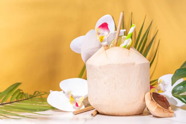 Young Thai coconut cocktail or juice, tropical summer drink in whole young coconut, with tropical leaves, decor and flowers