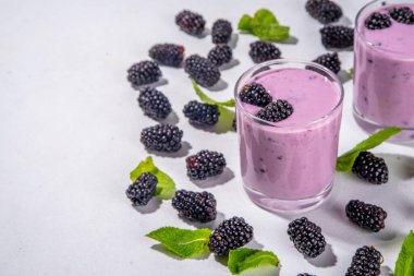İki bardak böğürtlenli smoothie, yoğurt veya taze böğürtlenli milkshake, beyaz masa arkasında nane yapraklı fotokopi alanı.