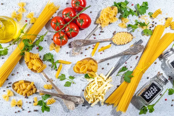 Beyaz masa arkasında çeşitli kurutulmuş spagetti, makarna ve kaşıklarla hazırlanmış makarna. Farklı şekillerde çiğ İtalyan makarnası, otlar, zeytinyağı, domatesler, baharatlar.