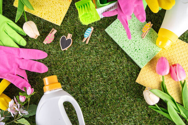 Весенняя уборка фона. Комплект чистящих средств, бутылок, аксессуаров и вещей с весенними цветами в корзине на белом деревянном зеленом саду