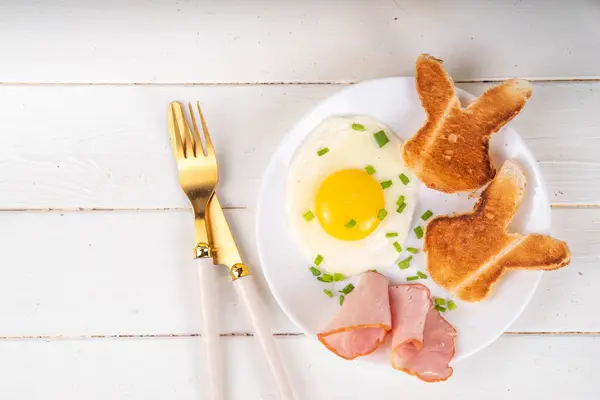 Easter breakfast or brunch with fried eggs, bacon slices and toasted bread shaped in form of bunny rabbits face, with spring flowers and Easter chocolate eggs on white wooden table copy space