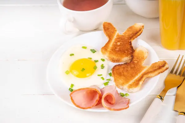 Easter breakfast or brunch with fried eggs, bacon slices and toasted bread shaped in form of bunny rabbits face, with spring flowers and Easter chocolate eggs on white wooden table copy space