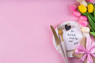 Anneler günü sofrası kurulumu. Pembe Anneler Günü 'nde kahvaltı, öğle yemeği, yemek menüsü, davetiye maketi. Altın çatal bıçak, tabak, lale çiçeği ve hediye kutusuyla güzel bir masa.