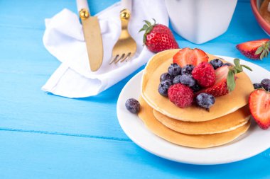 Yaz meyveli geleneksel Amerikan krepleri yığını. Sabah güneşinde bal, çilek ve yabanmersinli kahvaltı krepleri, mavi ahşap arka plan fotokopi alanı.