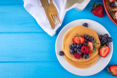 Yaz meyveli geleneksel Amerikan krepleri yığını. Sabah güneşinde bal, çilek ve yabanmersinli kahvaltı krepleri, mavi ahşap arka plan fotokopi alanı.