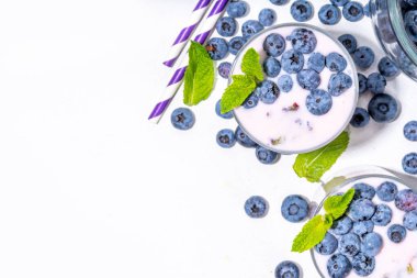 Üzerinde taze yaban mersini dekoru olan porsiyonlu bardaklarda yabanmersinli yoğurt, smoothie ya da milkshake ve arka planda beyaz mutfak masasında bir sürü taze böğürtlen.