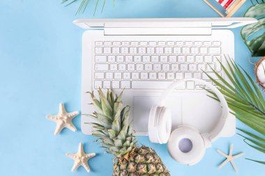 İş ve tatil. Yaratıcı yaz ofis dairesi beyaz dizüstü bilgisayarı, tropikal yaz aksesuarları, palmiye yaprakları, güneş gözlükleri, parmak arası terlikler, ananas ve açık mavi arka planda hindistan cevizi ile yatıyordu.