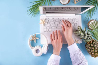 İş ve tatil. Yaratıcı yaz ofis dairesi beyaz dizüstü bilgisayarı, tropikal yaz aksesuarları, palmiye yaprakları, güneş gözlükleri, parmak arası terlikler, ananas ve açık mavi arka planda hindistan cevizi ile yatıyordu.