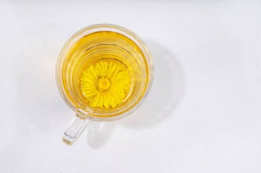 Karahindibalar bitki çayı eker. Şeffaf cam bardak ve çaydanlık, altın rengi çay içeceği, beyaz arka planda karahindiba çiçekleri ve yapraklarla.