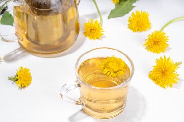 Karahindibalar bitki çayı eker. Şeffaf cam bardak ve çaydanlık, altın rengi çay içeceği, beyaz arka planda karahindiba çiçekleri ve yapraklarla.