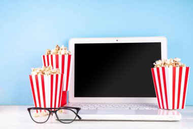 Sinema, film ve film izleme gecesi konsepti. Alkış tahtası, çizgili patlamış mısır kovası kutuları, açık mavi arka planda gözlüklü ve beyaz dizüstü bilgisayarlı, boşluğu kopyala