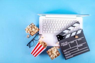 Sinema, film ve film izleme gecesi konsepti. Alkış tahtası, çizgili patlamış mısır kovası kutuları, açık mavi arka planda gözlüklü ve beyaz dizüstü bilgisayarlı, boşluğu kopyala