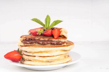 Stack of chocolate nutella filled pancakes. Sweet breakfast pancakes with chocolate sauce inside it, and fresh stawberries, on white kitchen table clipart