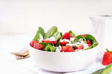 Ispanak ve çilekli yaz salatası süzme peynir ve balzamik sos taze çiğ sebze kasesi sağlıklı diyet yemeği beyaz mutfak masasında çatal bıçak takımı.