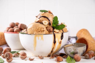 Fındıklı çikolatalı dondurma. Fındıklı dondurma kepçeli büyük kase porsiyonu, çikolata sosu ve mutfak masasının arkasında bir sürü fındık. 