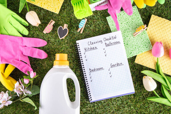Весенняя уборка фона. Комплект чистящих средств, бутылок, аксессуаров и вещей с весенними цветами в корзине на белом деревянном зеленом саду