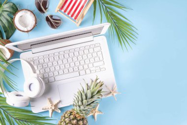 İş ve tatil. Yaratıcı yaz ofis dairesi beyaz dizüstü bilgisayarı, tropikal yaz aksesuarları, palmiye yaprakları, güneş gözlükleri, parmak arası terlikler, ananas ve açık mavi arka planda hindistan cevizi ile yatıyordu.