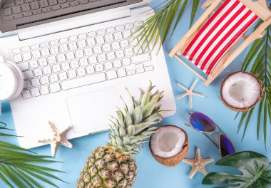 İş ve tatil. Yaratıcı yaz ofis dairesi beyaz dizüstü bilgisayarı, tropikal yaz aksesuarları, palmiye yaprakları, güneş gözlükleri, parmak arası terlikler, ananas ve açık mavi arka planda hindistan cevizi ile yatıyordu.